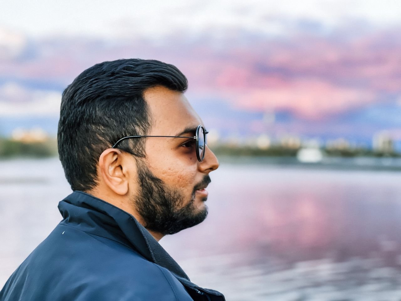 Bearded man in sunglasses gazing at sunset over tranquil lake, showcasing serene and thoughtful atmosphere.